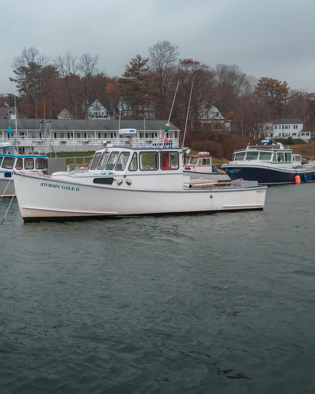 A quiet New England harbour on an overcast autumn day, with moored boats and white clapboard houses along the shore.
